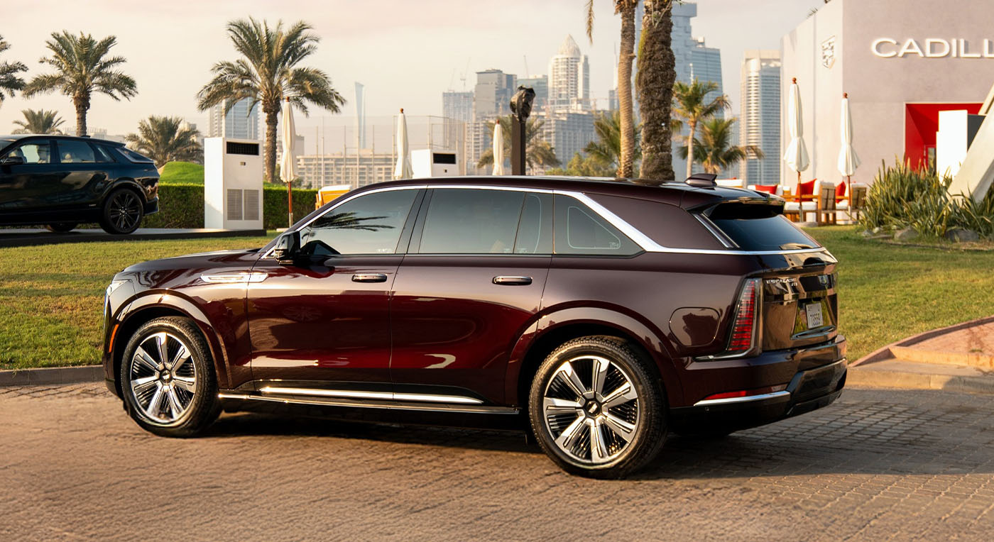 A sleek, Cadillac Escalade IQ dark red SUV is parked in a picturesque setting. Palm trees and a cityscape are in the background, evoking a sense of luxury and elegance.